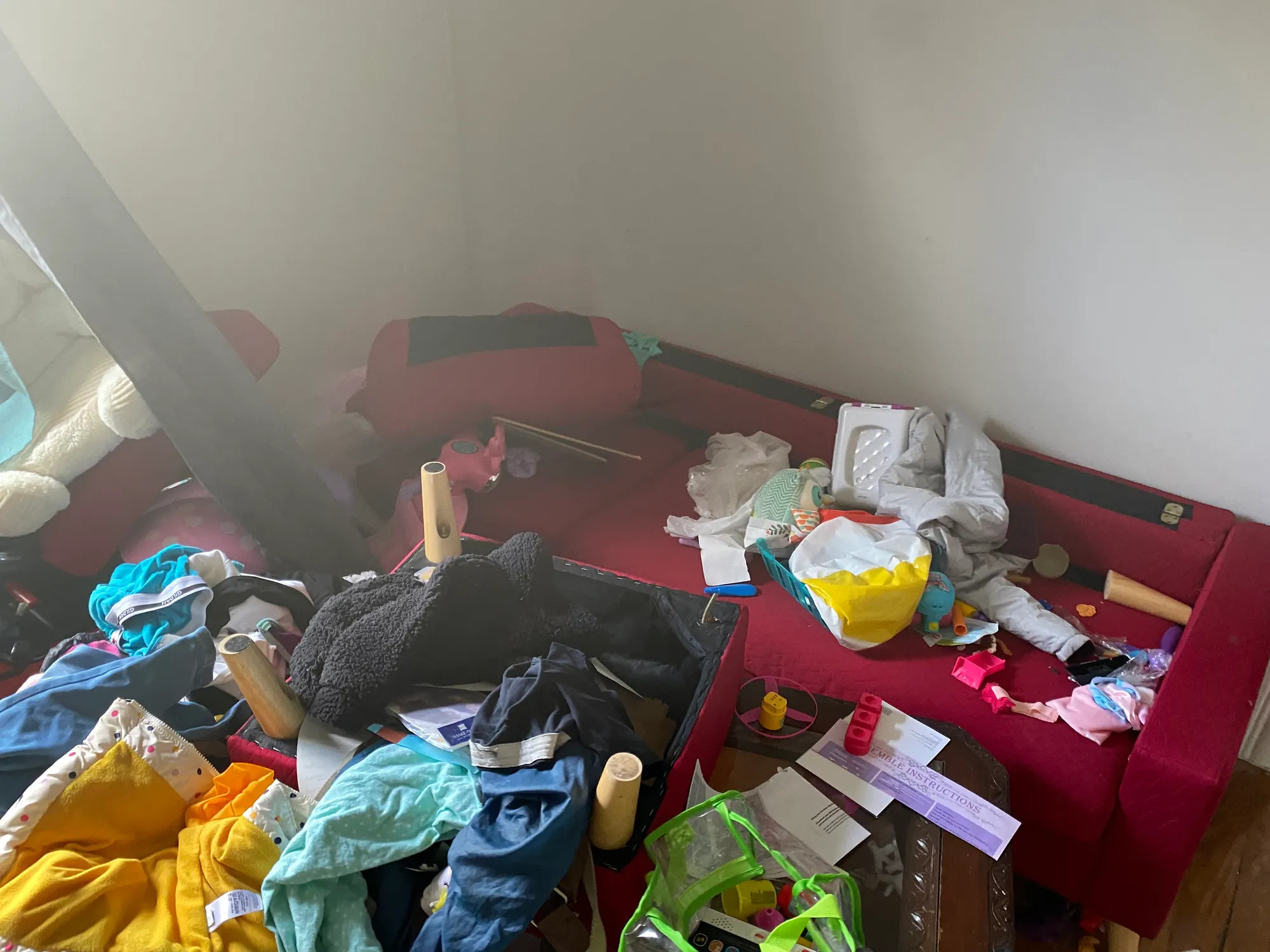 A cluttered room with a futon covered in clothing and toys.